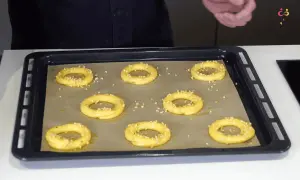 Huit ronds de pâte à choux, prêts pour devenir un délicieux Paris-Brest, saupoudrés de noisettes concassées sur une plaque de cuisson.
