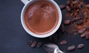 Chocolat chaud maison recette : une tasse fumante de chocolat chaud, entourée de fèves de cacao et de poudre de cacao. Une cuillère argentée repose à côté.
