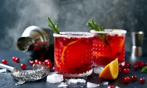 Deux verres de cocktail festif rouge vif, garni d'orange et de romarin, sur un fond sombre avec un shaker.