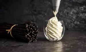 Une poche à douille dépose une crème diplomate vanillée dans un verre. Des gousses de vanille sont à côté, prêtes pour la recette.