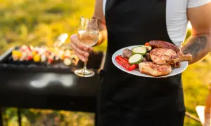 Un verre de vin blanc et une assiette de grillades, dont des côtelettes de porc et des saucisses, avec une dinde en arrière-plan sur le barbecue.
