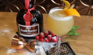 Un cocktail Grand Marnier, garni d'une écorce d'orange, à côté de la bouteille, d'un jigger en cuivre et de fausses baies rouges saupoudrées de sucre glace.
