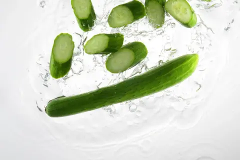 Tranches de concombre et un concombre entier tombant dans l'eau, parfait pour un cocktail concombre rafraîchissant.