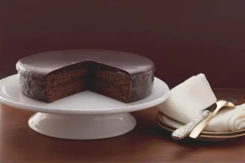 Un délicieux gâteau viennois au chocolat, nappé d'un glaçage brillant, est présenté sur un présentoir blanc. Une part est retirée, révélant sa texture moelleuse.