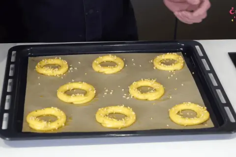 Huit ronds de pâte à choux, prêts pour devenir un délicieux Paris-Brest, saupoudrés de noisettes concassées sur une plaque de cuisson.