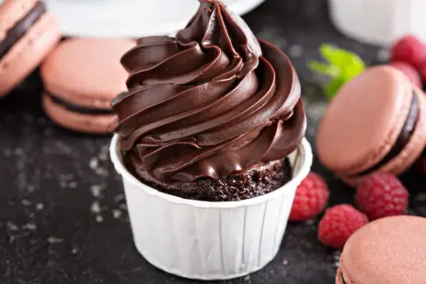 Un cupcake au chocolat surmonté d'une ganache montée chocolat au lait onctueuse, entouré de macarons et de framboises.
