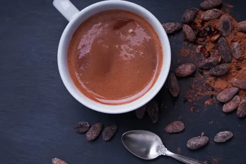 Chocolat chaud maison recette : une tasse fumante de chocolat chaud, entourée de fèves de cacao et de poudre de cacao. Une cuillère argentée repose à côté.