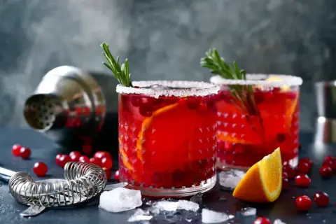 Deux verres de cocktail festif rouge vif, garni d'orange et de romarin, sur un fond sombre avec un shaker.