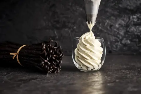 Une poche à douille dépose une crème diplomate vanillée dans un verre. Des gousses de vanille sont à côté, prêtes pour la recette.