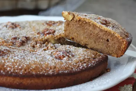 Gâteau aux noix, saupoudré de sucre glace. Une part est soulevée, révélant la texture moelleuse de cette recette de noix.