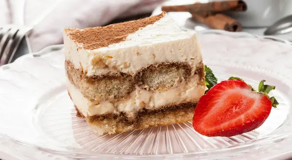 Tranche de tiramisu sans oeuf, garnie de cacao et d'une fraise fraîche, sur une assiette rose.