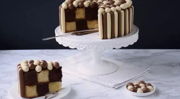 Un gâteau damier, décoré de bâtonnets et de billes chocolatées, présente une tranche coupée révélant ses couches alternées.