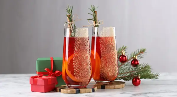 Deux verres de cocktail de Noël facile et rapide, décorés d'orange et de cannelle, avec des cadeaux et un sapin en arrière-plan.
