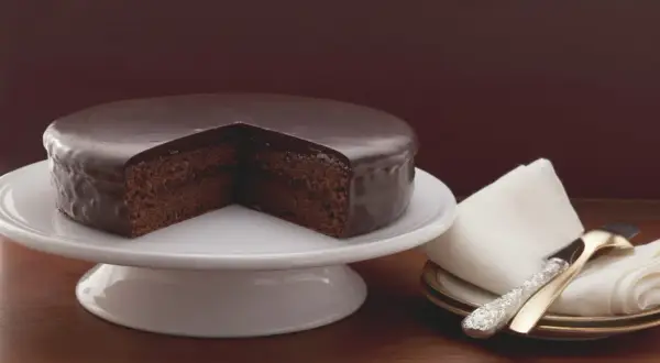 Un délicieux gâteau viennois au chocolat, nappé d'un glaçage brillant, est présenté sur un présentoir blanc. Une part est retirée, révélant sa texture moelleuse.