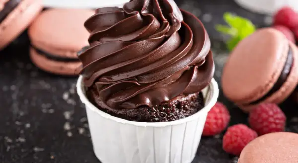 Un cupcake au chocolat surmonté d'une ganache montée chocolat au lait onctueuse, entouré de macarons et de framboises.