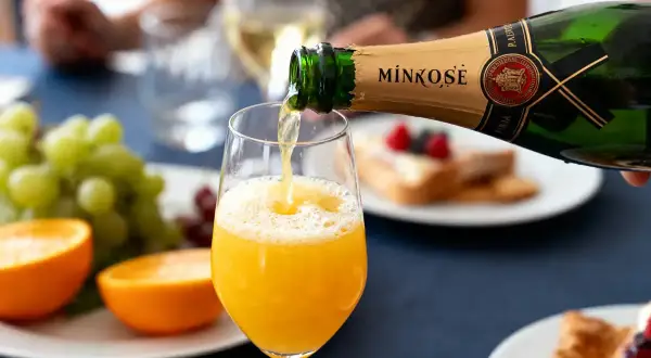 Une bouteille de Mimosé verse son contenu pétillant dans un verre rempli de jus d'orange, créant un cocktail mimosa parfait pour un brunch.