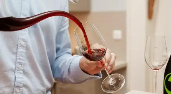 Un homme verse du vin rouge d'un **decanter ou carafer** dans un verre. Une autre bouteille et un verre vide sont visibles.