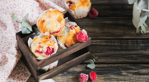 Muffins framboise dorés et moelleux dans une caisse en bois, décorés de quelques framboises fraîches.