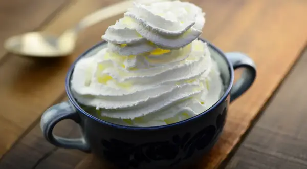 Une tasse de chantilly maison, aérienne et onctueuse, prête à être dégustée. Une cuillère dorée est posée à côté.
