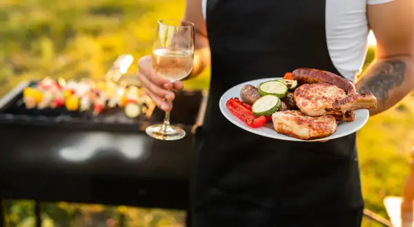 Un verre de vin blanc et une assiette de grillades, dont des côtelettes de porc et des saucisses, avec une dinde en arrière-plan sur le barbecue.