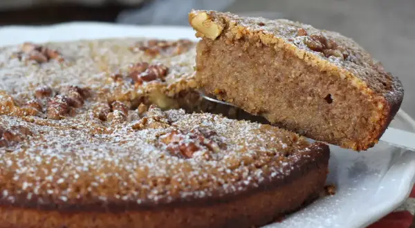 Gâteau aux noix, saupoudré de sucre glace. Une part est soulevée, révélant la texture moelleuse de cette recette de noix.
