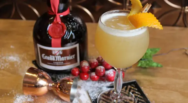 Un cocktail Grand Marnier, garni d'une écorce d'orange, à côté de la bouteille, d'un jigger en cuivre et de fausses baies rouges saupoudrées de sucre glace.