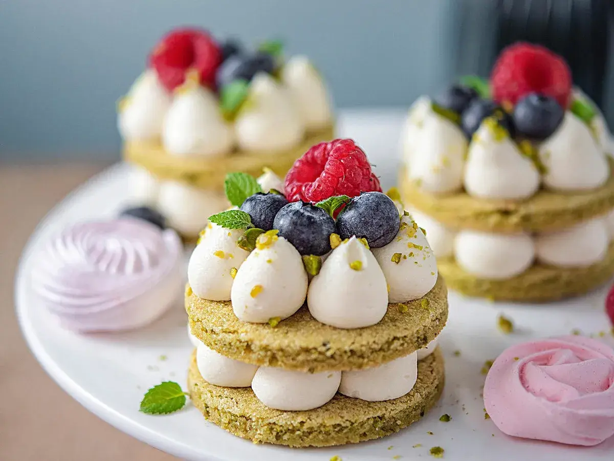 Petits gâteaux verts garnis de ganache montée chocolat blanc facile, fruits rouges et pistaches. Meringues roses en arrière-plan.