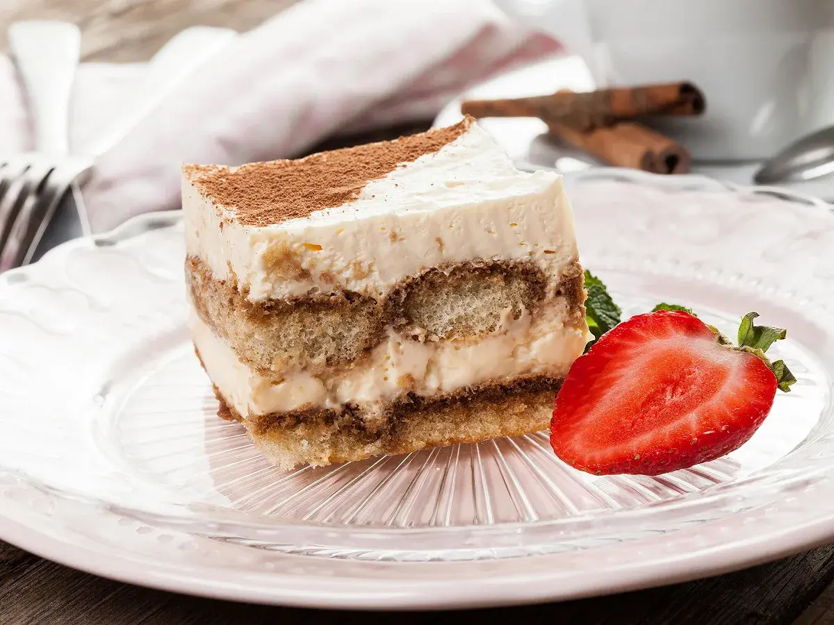 Tranche de tiramisu sans oeuf, garnie de cacao et d'une fraise fraîche, sur une assiette rose.