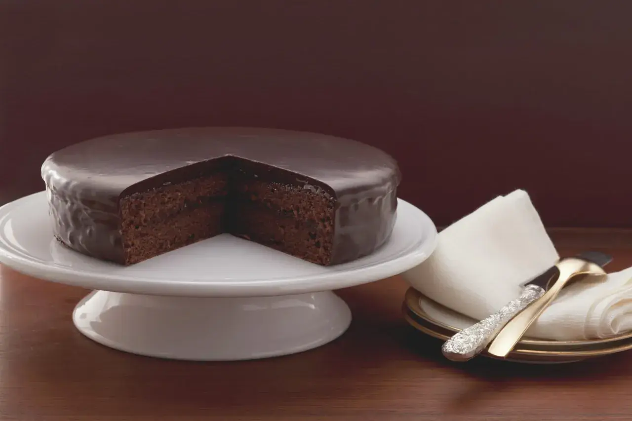 Un délicieux gâteau viennois au chocolat, nappé d'un glaçage brillant, est présenté sur un présentoir blanc. Une part est retirée, révélant sa texture moelleuse.