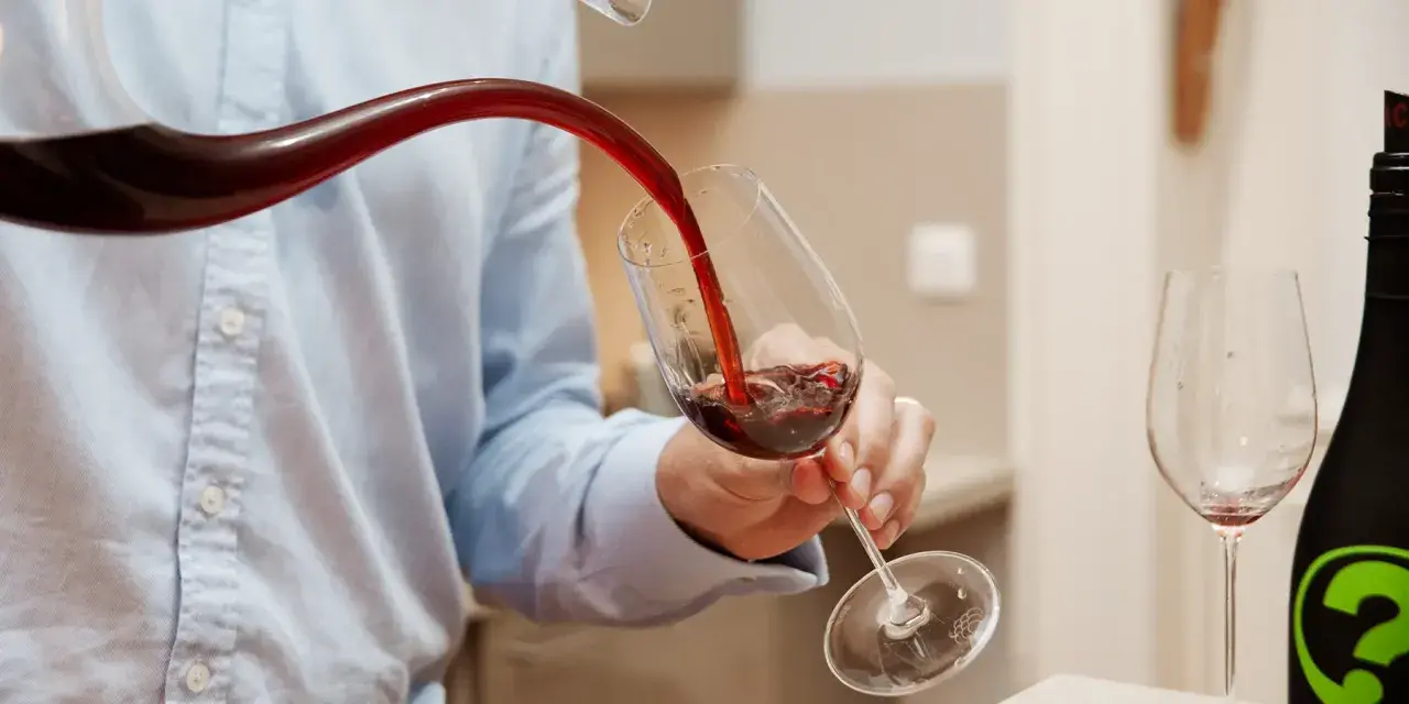 Un homme verse du vin rouge d'un **decanter ou carafer** dans un verre. Une autre bouteille et un verre vide sont visibles.
