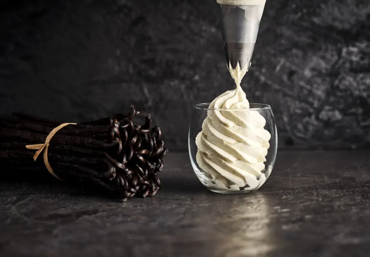 Une poche à douille dépose une crème diplomate vanillée dans un verre. Des gousses de vanille sont à côté, prêtes pour la recette.