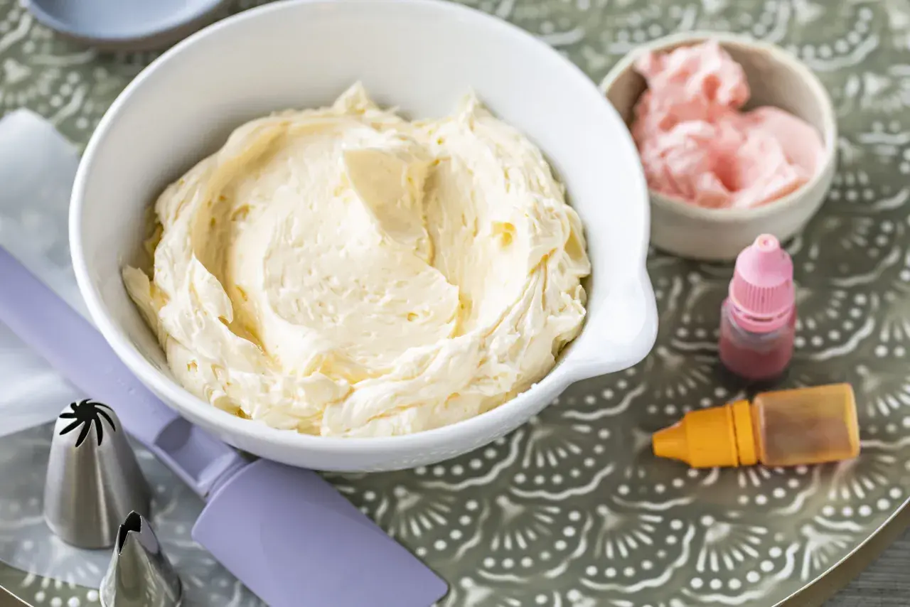 Crème au beurre à la meringue suisse onctueuse dans un bol blanc, avec des colorants et des douilles de pâtisserie.