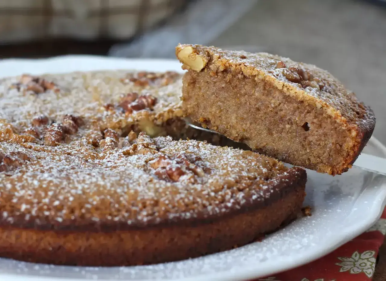 Gâteau aux noix, saupoudré de sucre glace. Une part est soulevée, révélant la texture moelleuse de cette recette de noix.