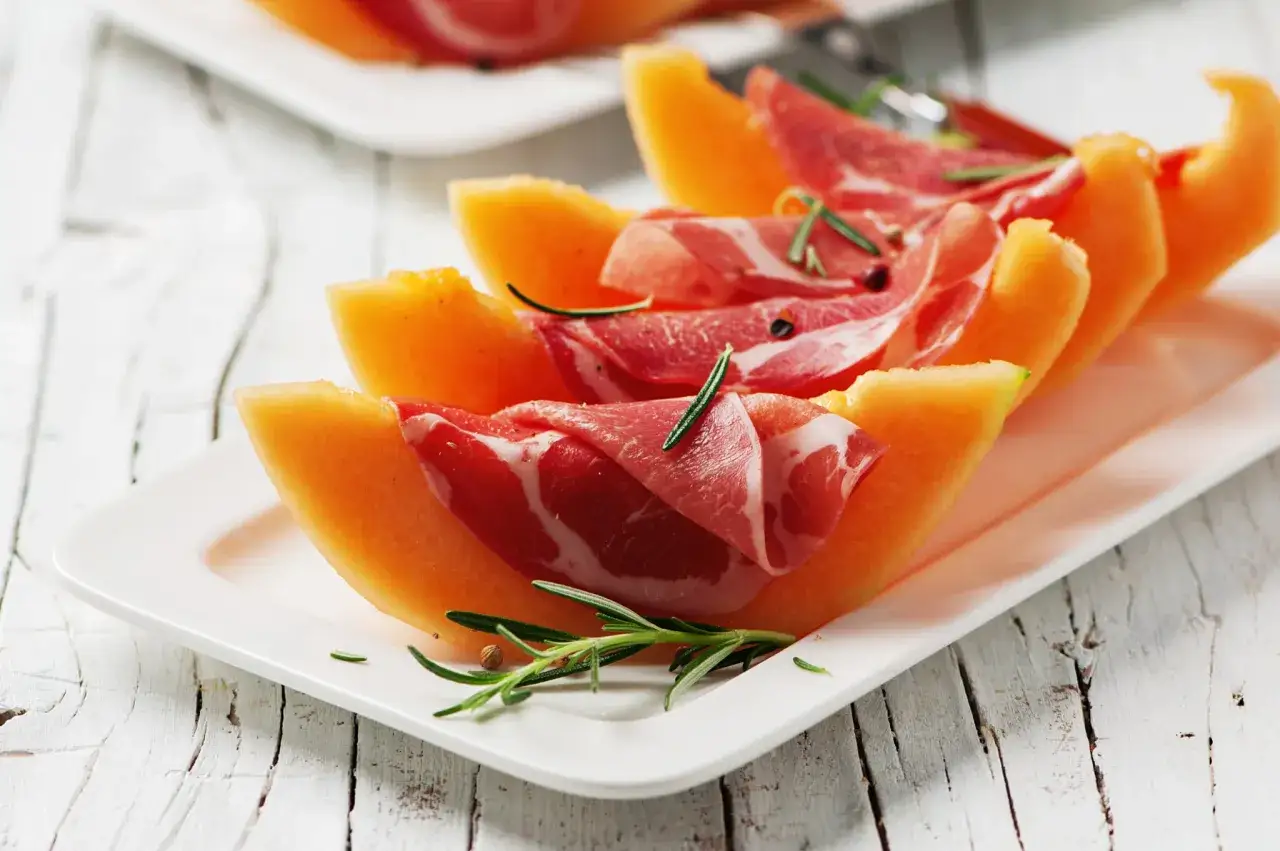 Tranches de melon et jambon cru sur une assiette blanche, garnies de romarin. Une idée parfaite pour savoir quel vin avec melon et jambon cru.