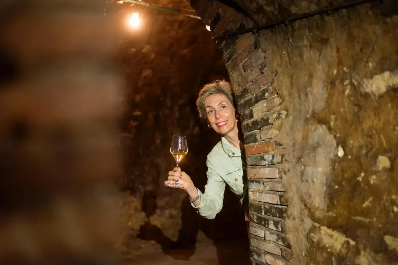 Une femme souriante tient un verre de vin clair dans une cave voûtée en briques.