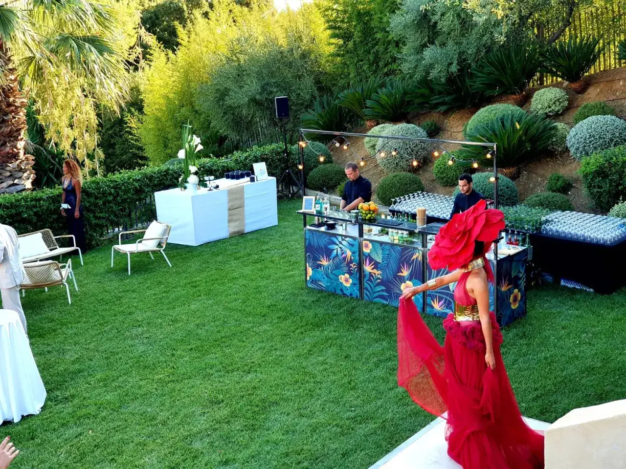 Une femme en robe rouge spectaculaire se tient près d'un bar tropical, prête pour un cocktail mariage. Des barmans préparent des boissons dans un jardin luxuriant.