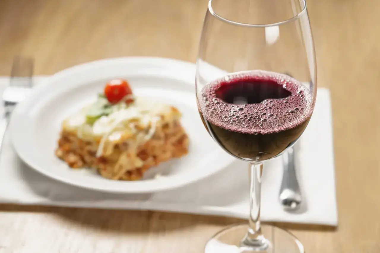 Une part de lasagne gratinée, garnie d'une tomate cerise, est servie à côté d'un verre de vin rouge pétillant.