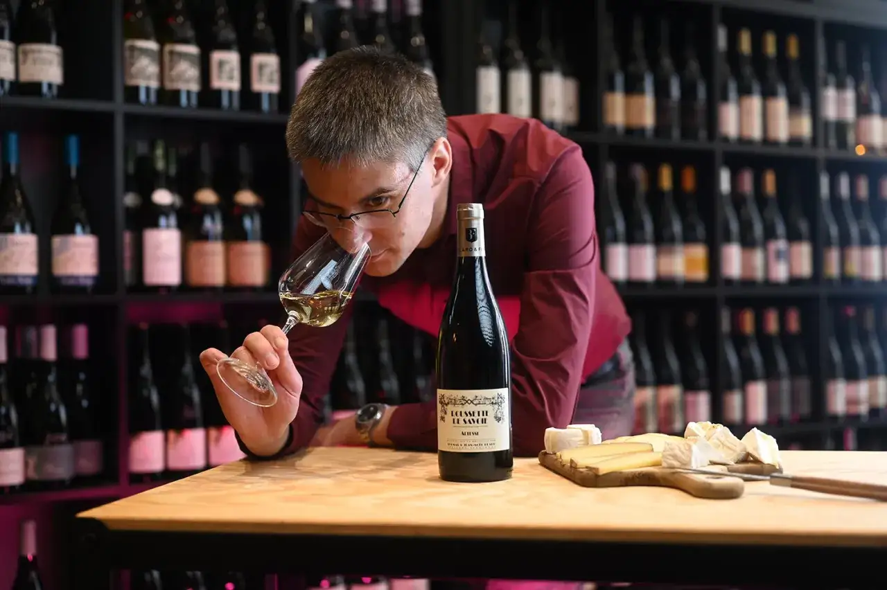 Un homme déguste un vin blanc sec, peut-être un des top 10, devant une étagère remplie de bouteilles. Fromage et pain sont sur la table.