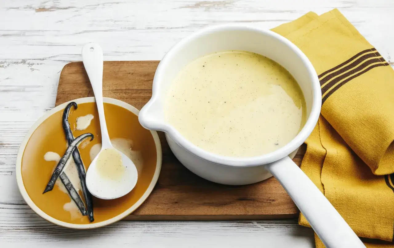 Une casserole de crème anglaise traditionnelle, avec des gousses de vanille sur une assiette et une cuillère.