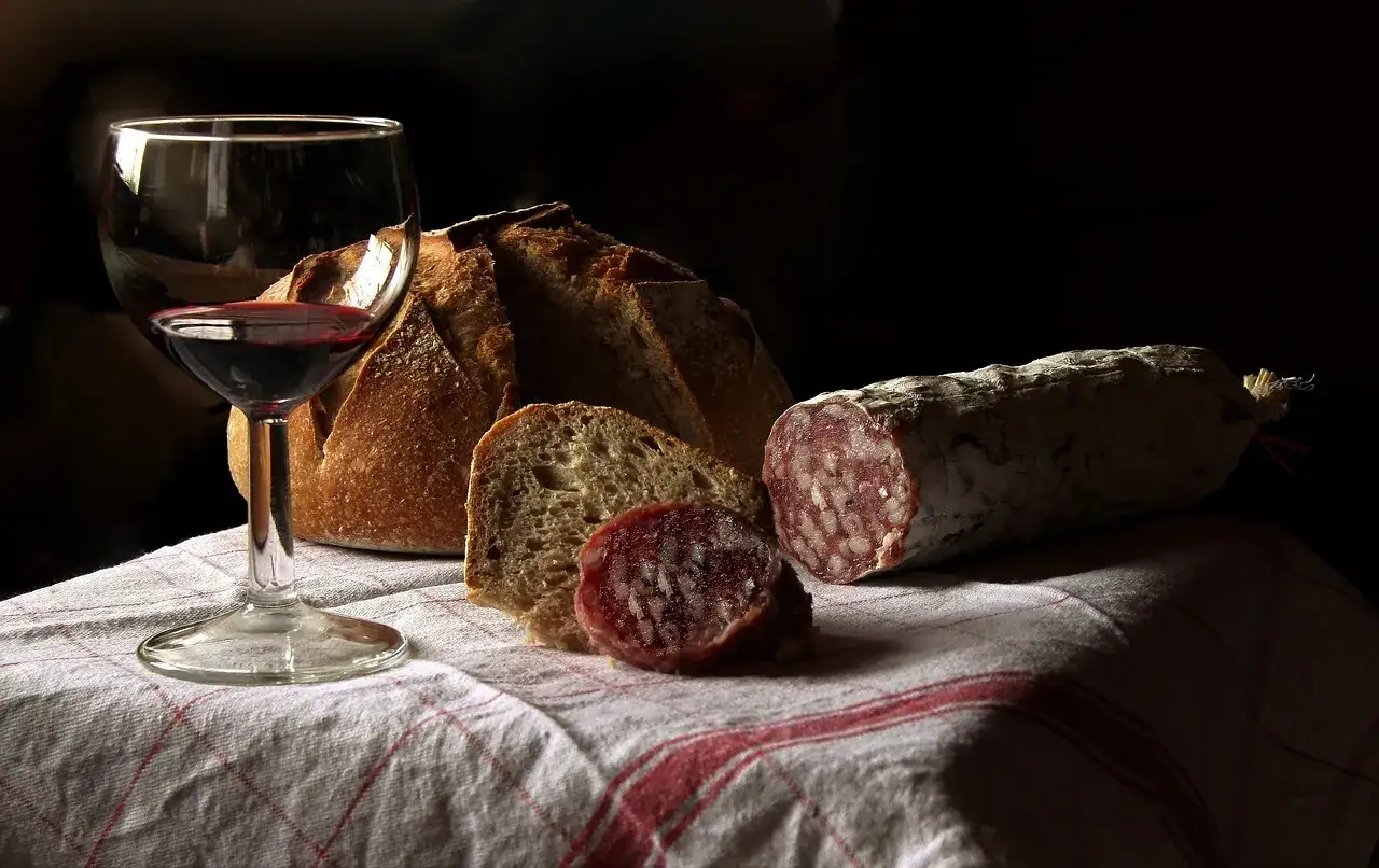 Un verre de vin rouge apéritif, du pain artisanal et un saucisson sec sur une nappe à carreaux rouges.