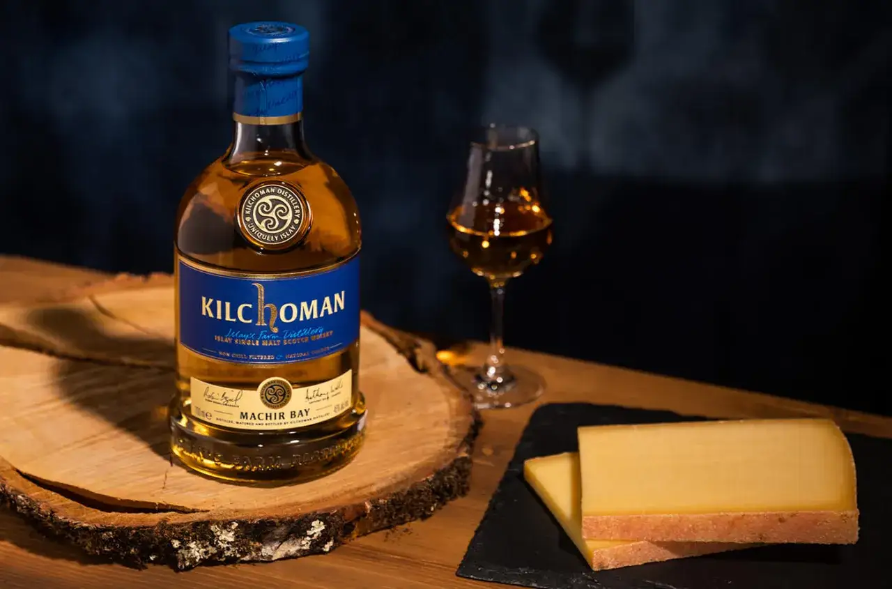 Bouteille de whisky Kilchoman Machir Bay et un verre de whisky, parfaits pour un digestif. Des tranches de fromage complètent la scène.