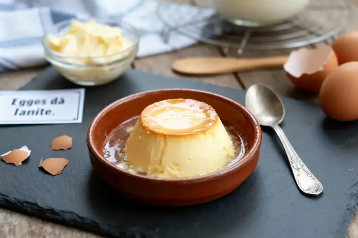 Un délicieux oeuf au lait, onctueux et doré, servi dans un ramequin en terre cuite. Des coquilles d'oeufs et une cuillère complètent la scène.