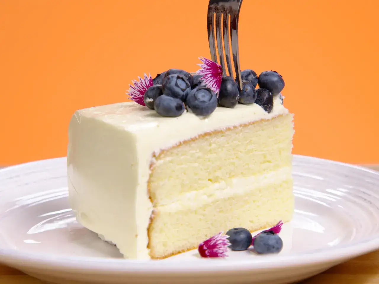 Tranche de gâteau moelleux, nappée d'une onctueuse crème au beurre, décorée de myrtilles et de fleurs. Une fourchette s'apprête à déguster ce délice.