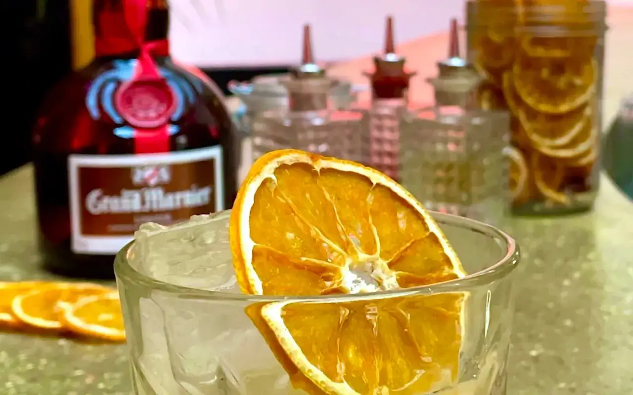 Un verre rempli de glaçons et d'une tranche d'orange séchée, avec une bouteille de Grand Marnier en arrière-plan. Un cocktail parfait pour se détendre.
