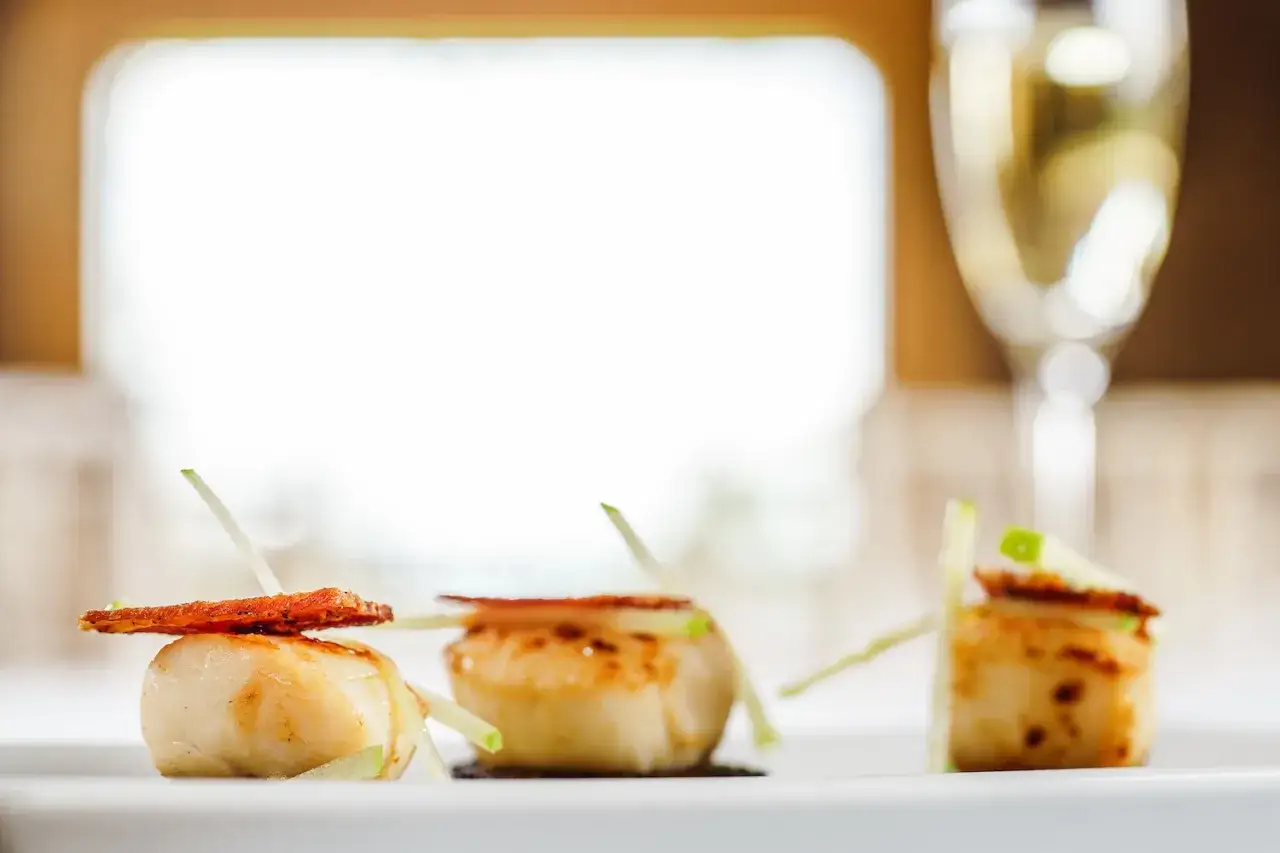 Trois noix de Saint-Jacques poêlées, garnies d'une fine tranche de carotte et de bâtonnets de pomme verte. Un verre de vin blanc pétillant suggère quel vin avec coquille Saint-Jacques.