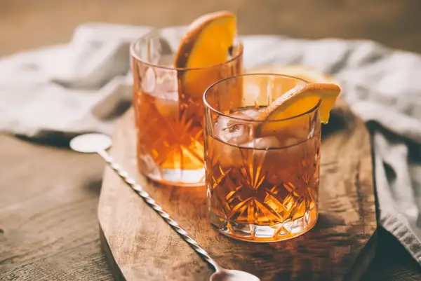 Deux verres d'armagnac digestif ambré, glacés et garnis d'une tranche d'orange, sur une planche en bois.