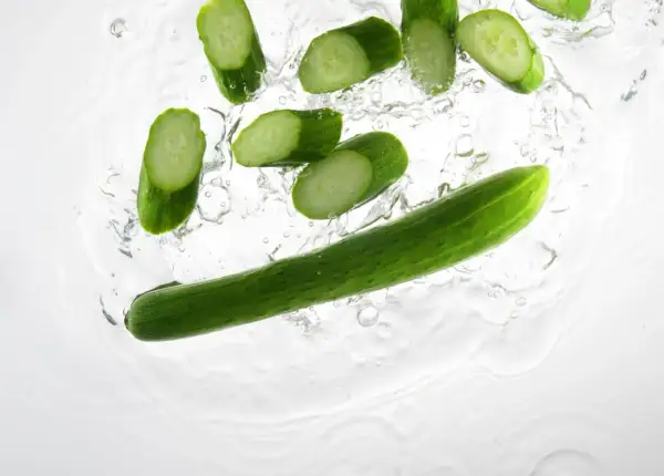Tranches de concombre et un concombre entier tombant dans l'eau, parfait pour un cocktail concombre rafraîchissant.