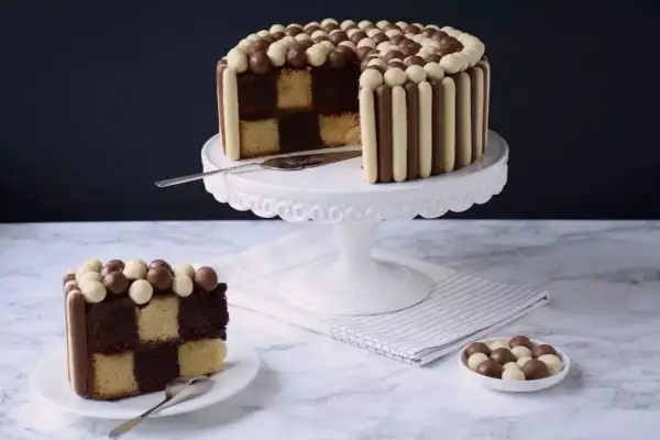 Un gâteau damier, décoré de bâtonnets et de billes chocolatées, présente une tranche coupée révélant ses couches alternées.