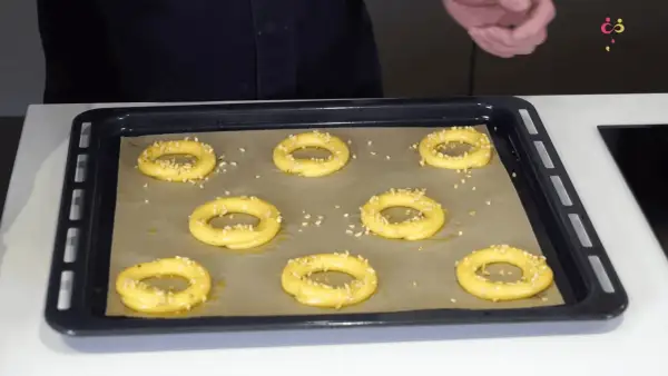 Huit ronds de pâte à choux, prêts pour devenir un délicieux Paris-Brest, saupoudrés de noisettes concassées sur une plaque de cuisson.