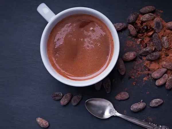 Chocolat chaud maison recette : une tasse fumante de chocolat chaud, entourée de fèves de cacao et de poudre de cacao. Une cuillère argentée repose à côté.